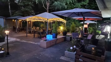 An outdoor patio area with tables and chairs under umbrellas, surrounded by potted plants and trees.