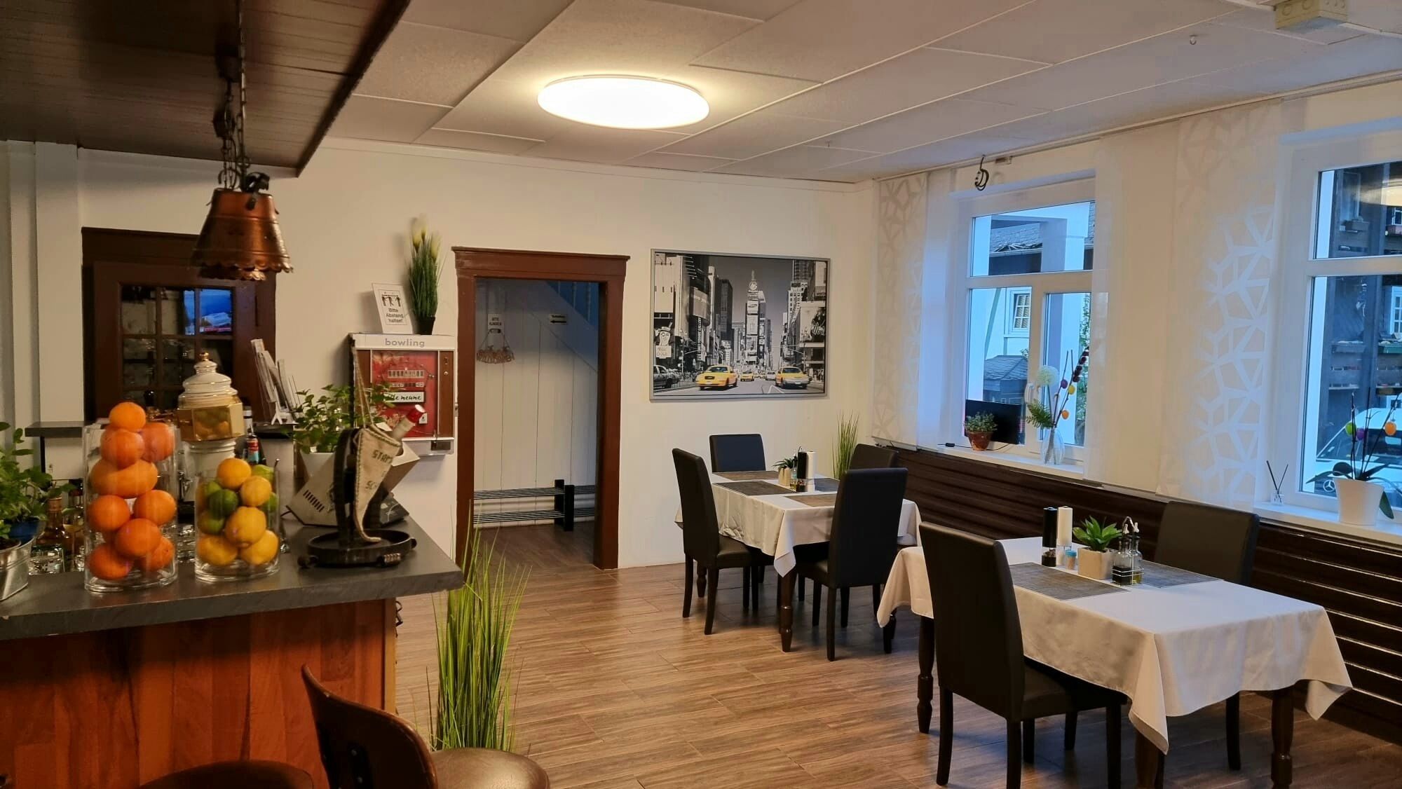 Un restaurante con piso de madera, una zona de bar con frutas y botellas, y una pintura de paisaje urbano en la pared.