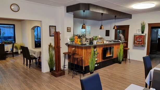 Ein moderner Barbereich mit einer Holztheke, Barhocker und einem Kamin. Es gibt Blumentöpfe und eine Uhr an der Wand.