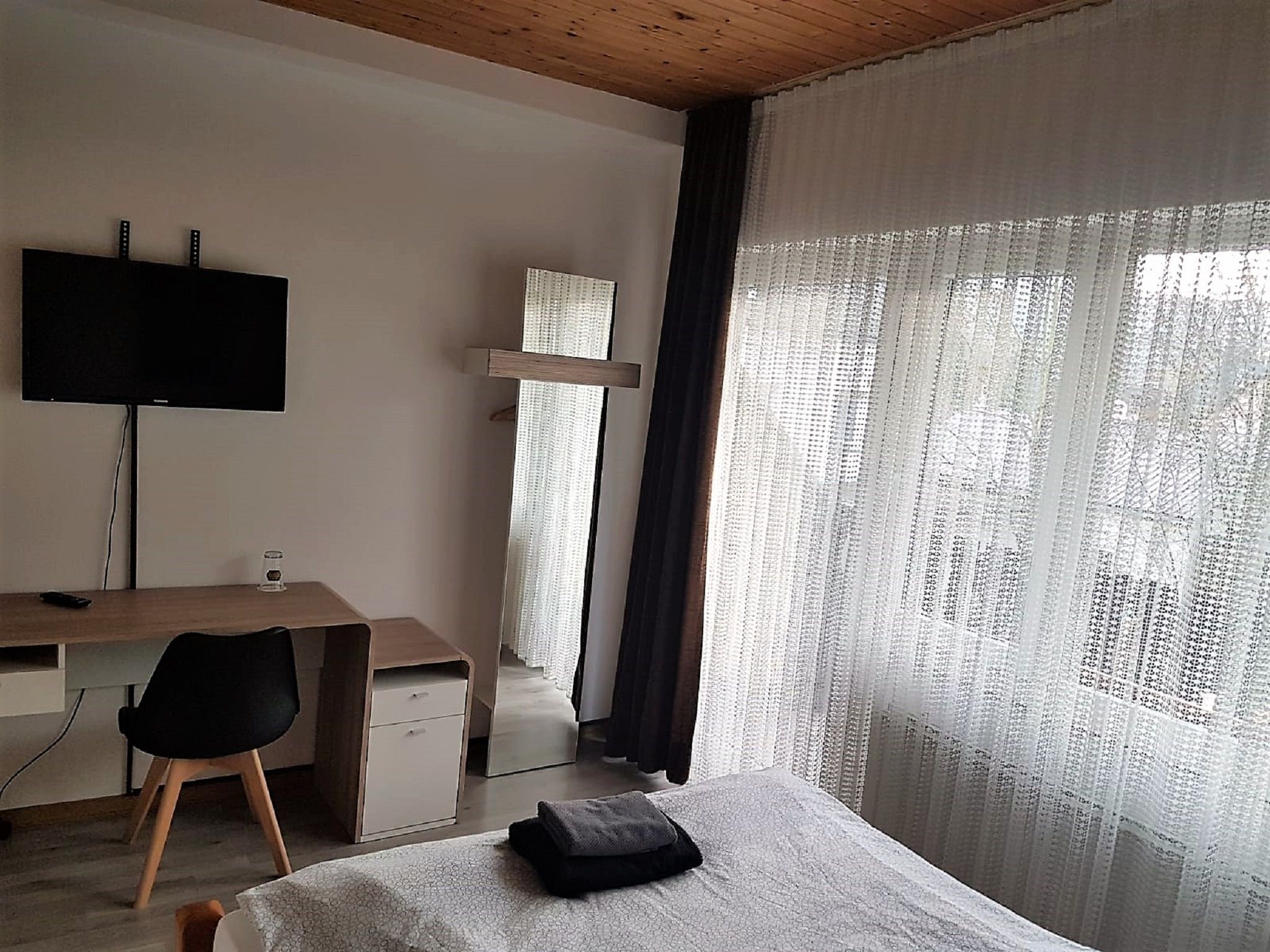 A bedroom with a bed, a desk, a chair, a TV, and a window with sheer curtains.
