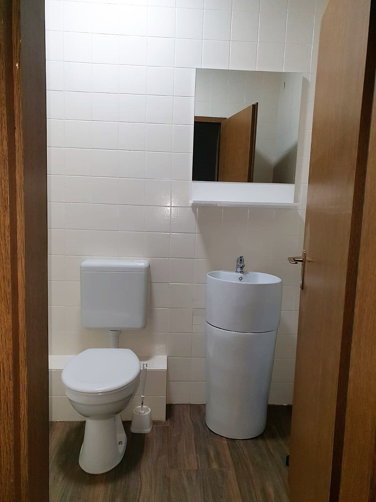 Un baño pequeño y limpio con azulejos blancos, un inodoro, un lavabo y un espejo sobre el lavabo.