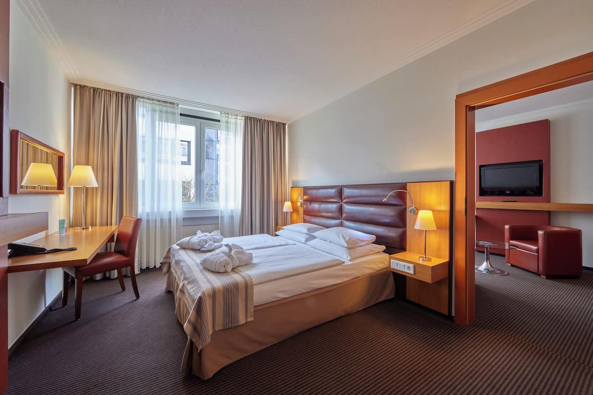 A hotel room with a large bed, two bedside tables with lamps, a desk with a chair, a TV, and a red armchair.
