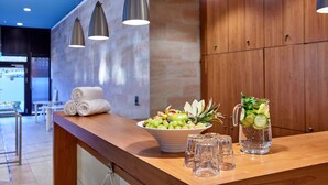 Un espace d’accueil d’hôtel avec un comptoir en bois, un saladier rempli de fruits, une carafe de thé glacé et des serviettes enroulées.
