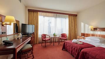 Habitación de hotel con cama, escritorio con computadora, televisor, dos sillas, una mesita y ventana con cortinas.