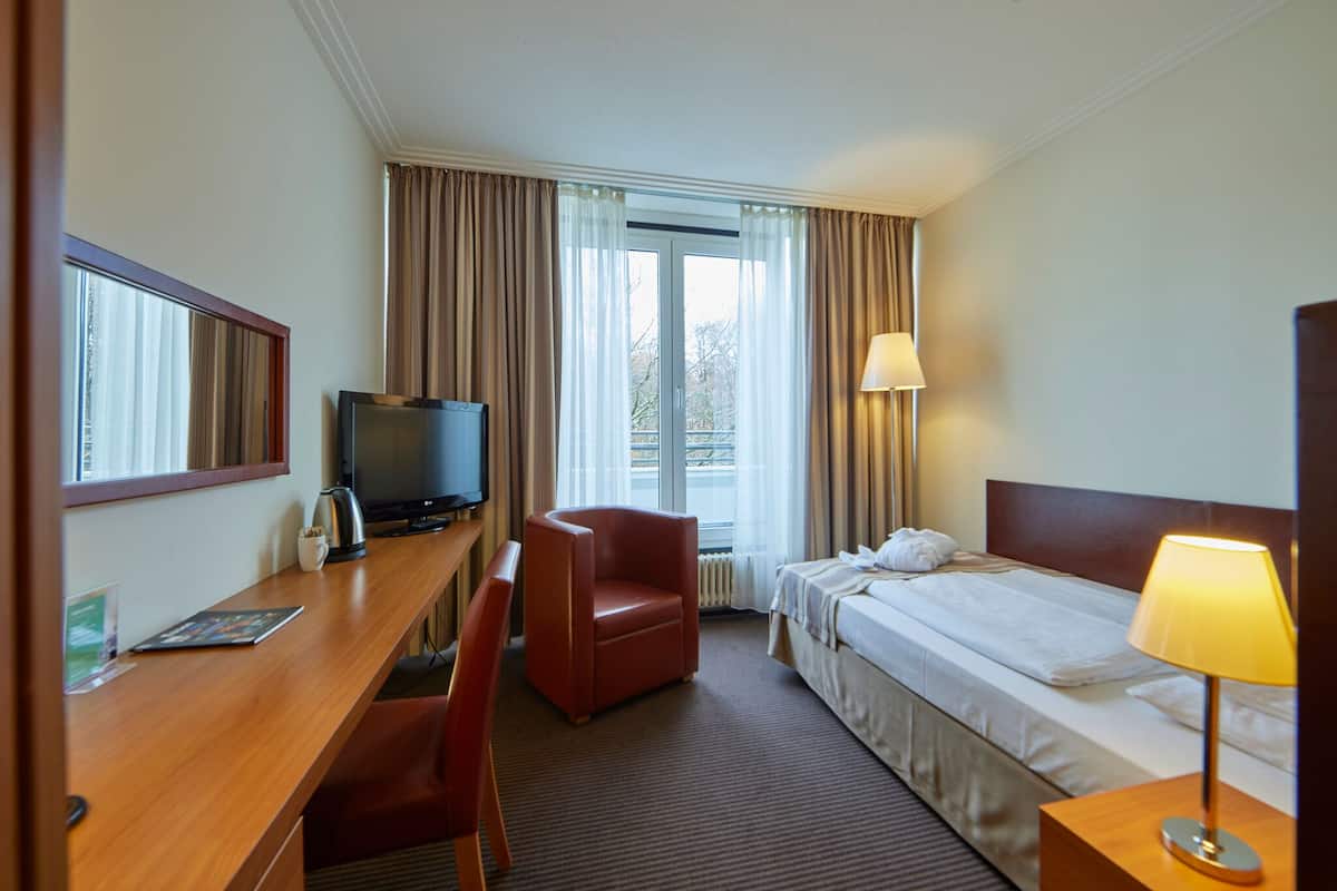 A hotel room with a bed, a desk with a television, a chair, a lamp, and a window with curtains.