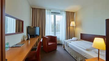 Ein Hotelzimmer mit einem Bett, einem Schreibtisch mit Fernseher, einem Sessel, einer Lampe und einem Fenster mit Vorhängen.