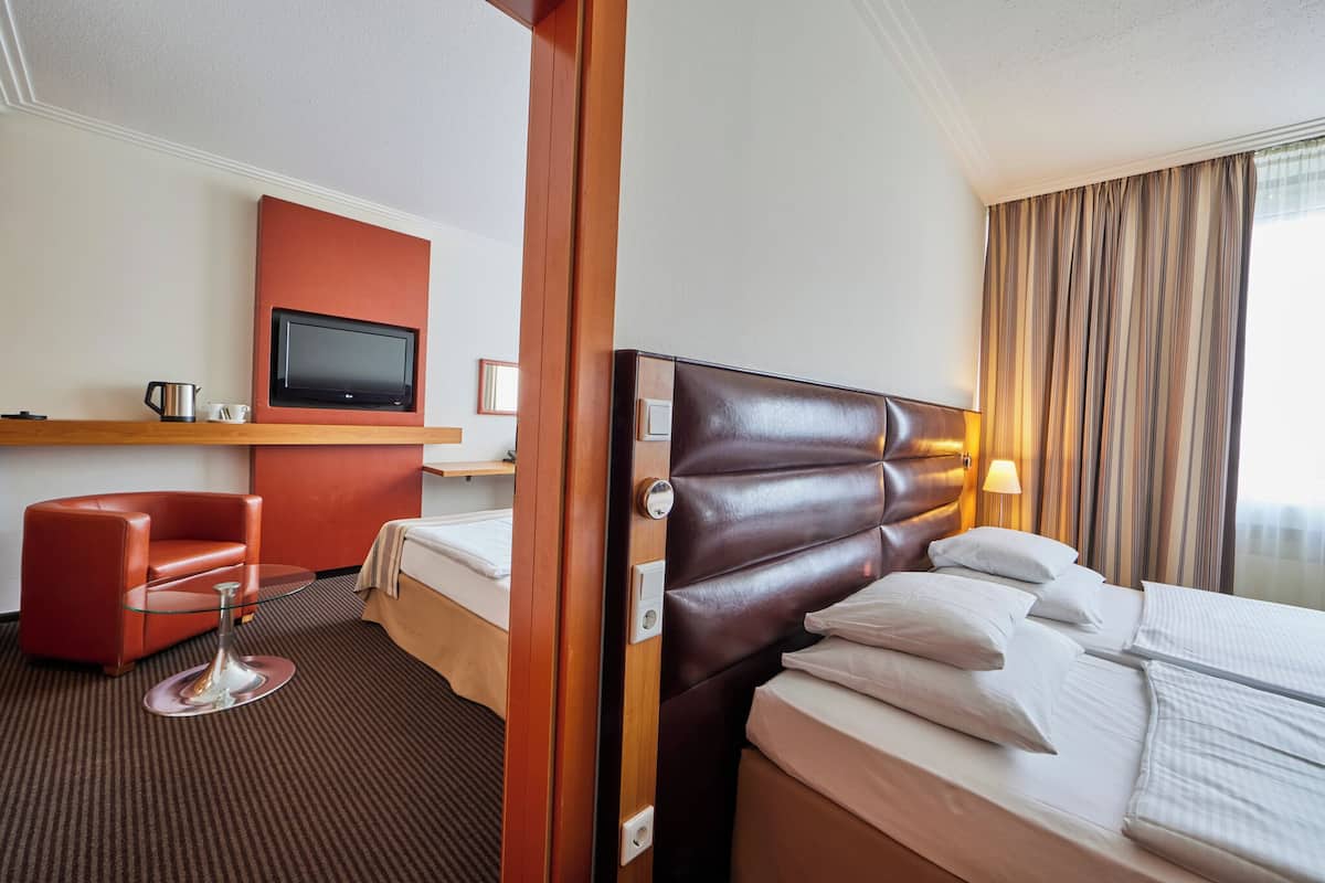 A hotel room with a bed, a TV mounted on the wall, a red armchair, and a small table with a lamp.