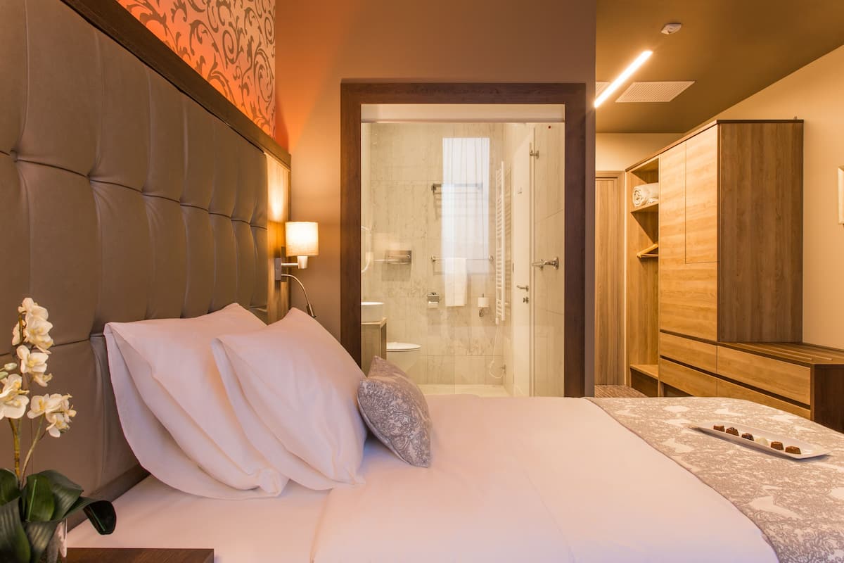 Une chambre d’hôtel avec un grand lit, une armoire en bois, une salle de bain avec une douche en verre et une petite table sur laquelle est posé un vase de fleurs.