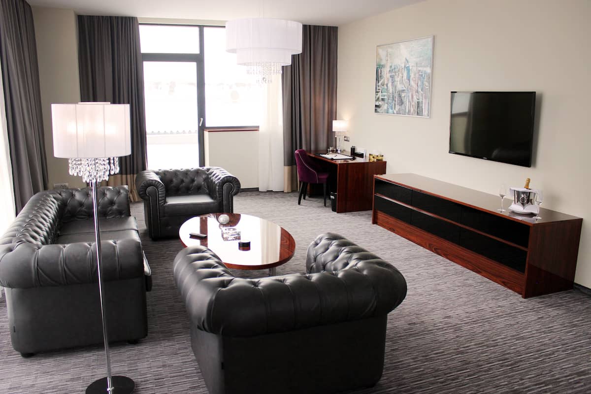 Une chambre d’hôtel moderne dotée d’un espace salon comprenant un canapé, une table basse, un bureau avec une chaise, une télévision et une grande fenêtre avec des rideaux.