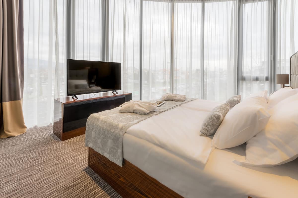 Une chambre d’hôtel avec un grand lit, un téléviseur à écran plat, une table de chevet en bois et une vue sur la ville à travers des rideaux transparents.