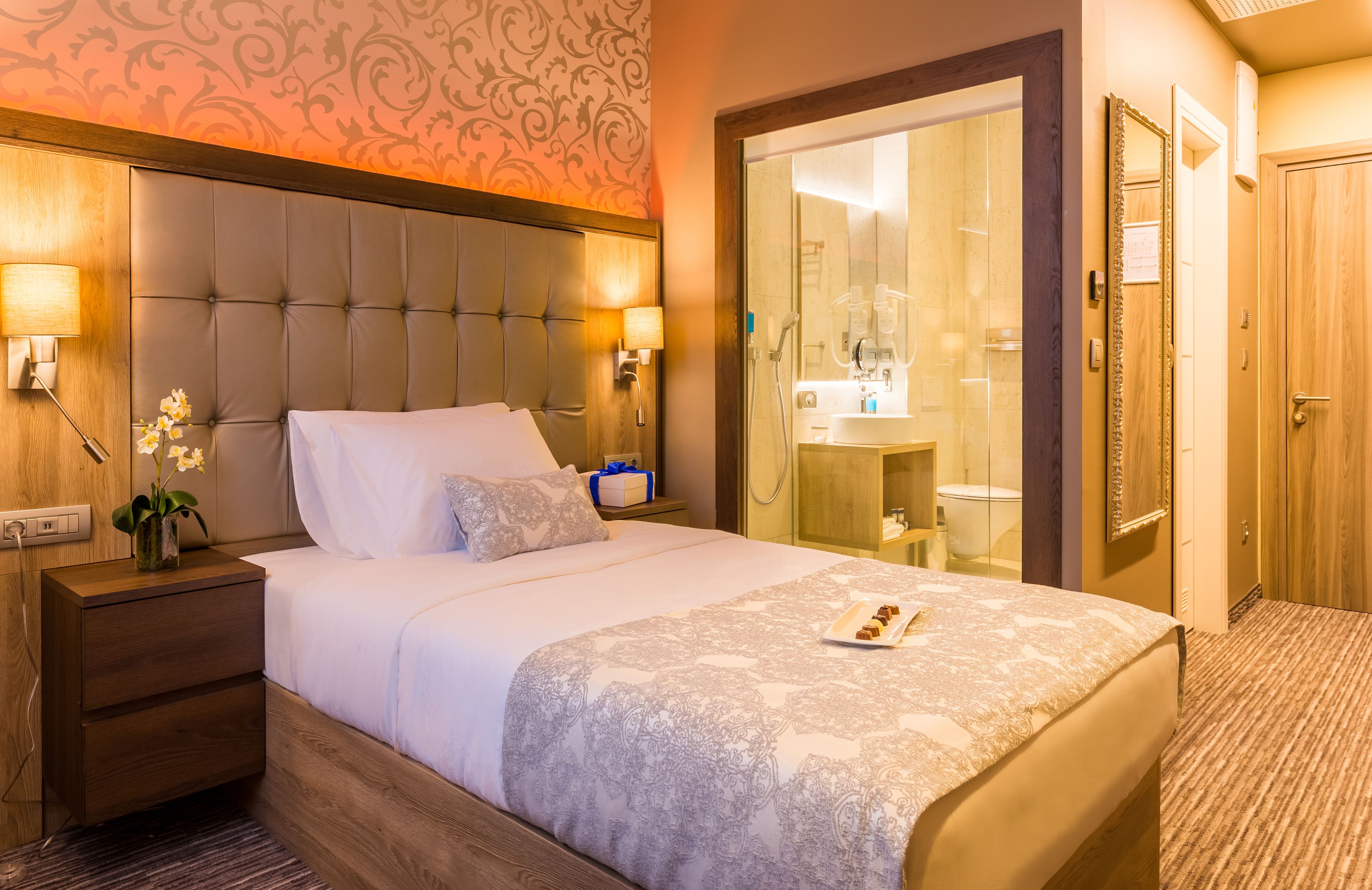 A hotel room with a large bed, bedside lamps, a nightstand, a vase with flowers, and a bathroom visible through a glass door.