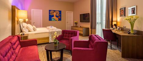 Quarto de hotel com uma cama, duas poltronas vermelhas, uma mesinha com flores, uma escrivaninha com cadeira, uma televisão e obras de arte nas paredes.