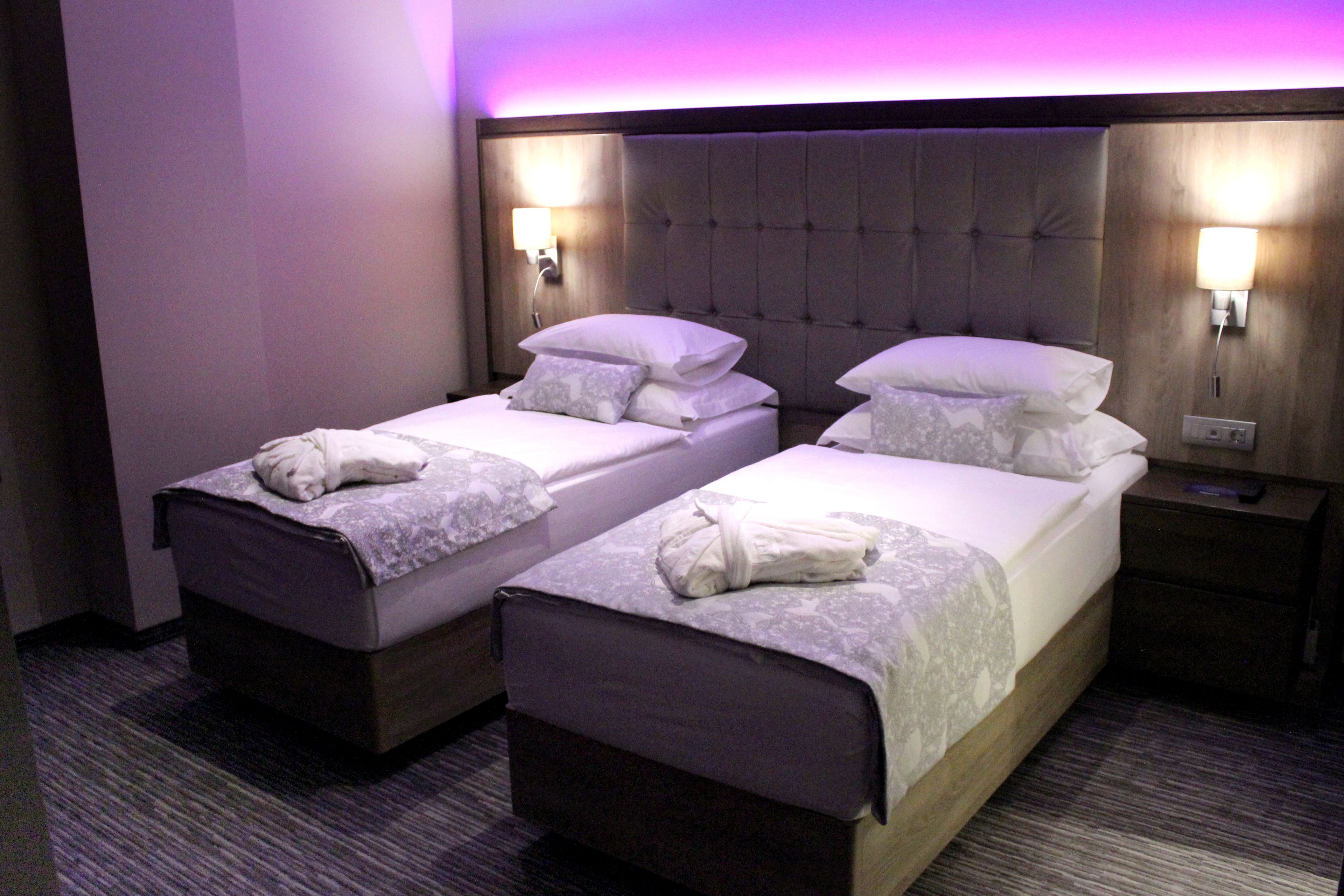 A hotel room with two single beds, each with a pillow and a folded towel. There are two wall-mounted lamps on either side of the beds.