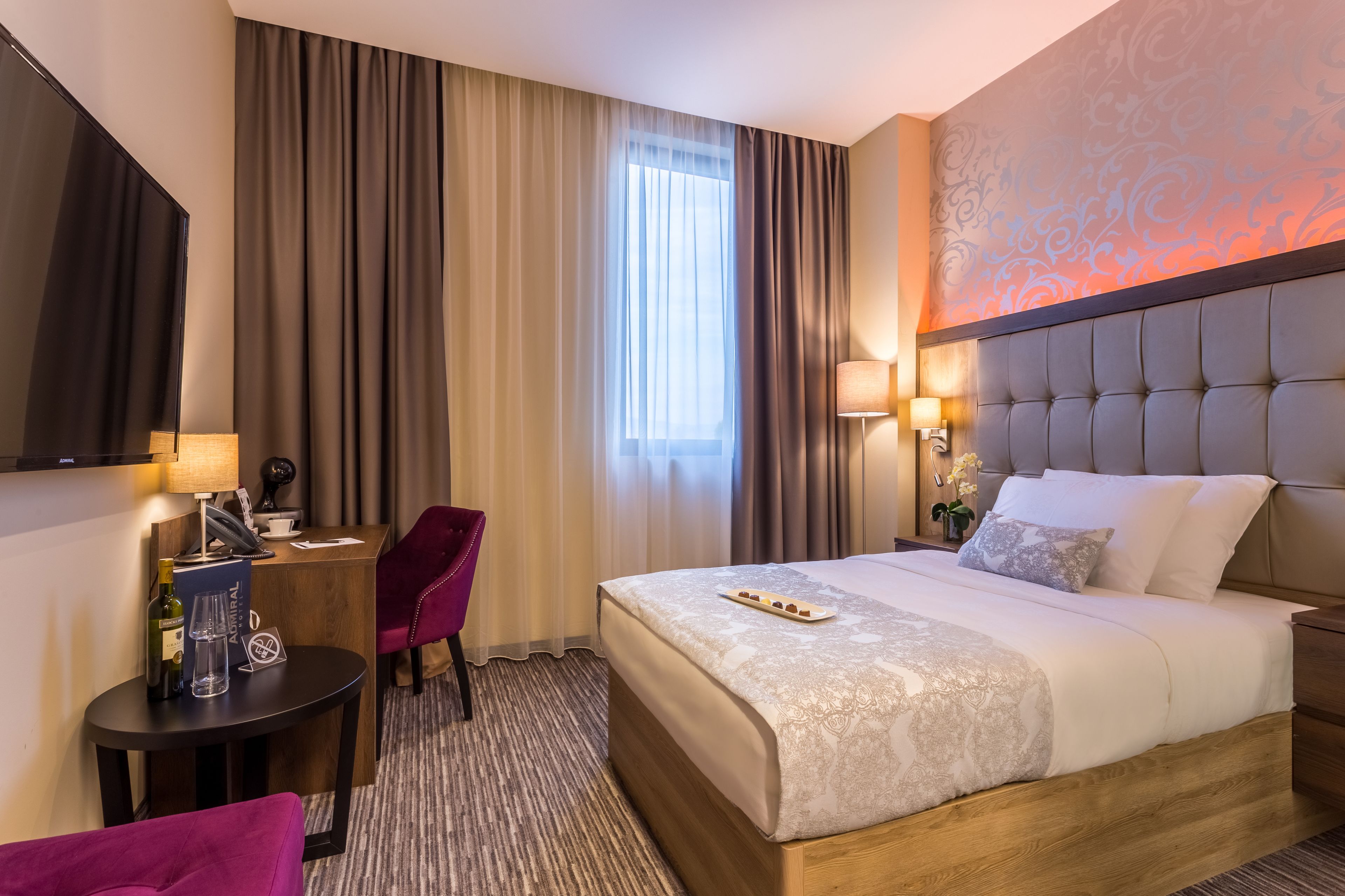 Una camera d'albergo con un letto grande, una scrivania con una sedia, una TV e una finestra con tende.
