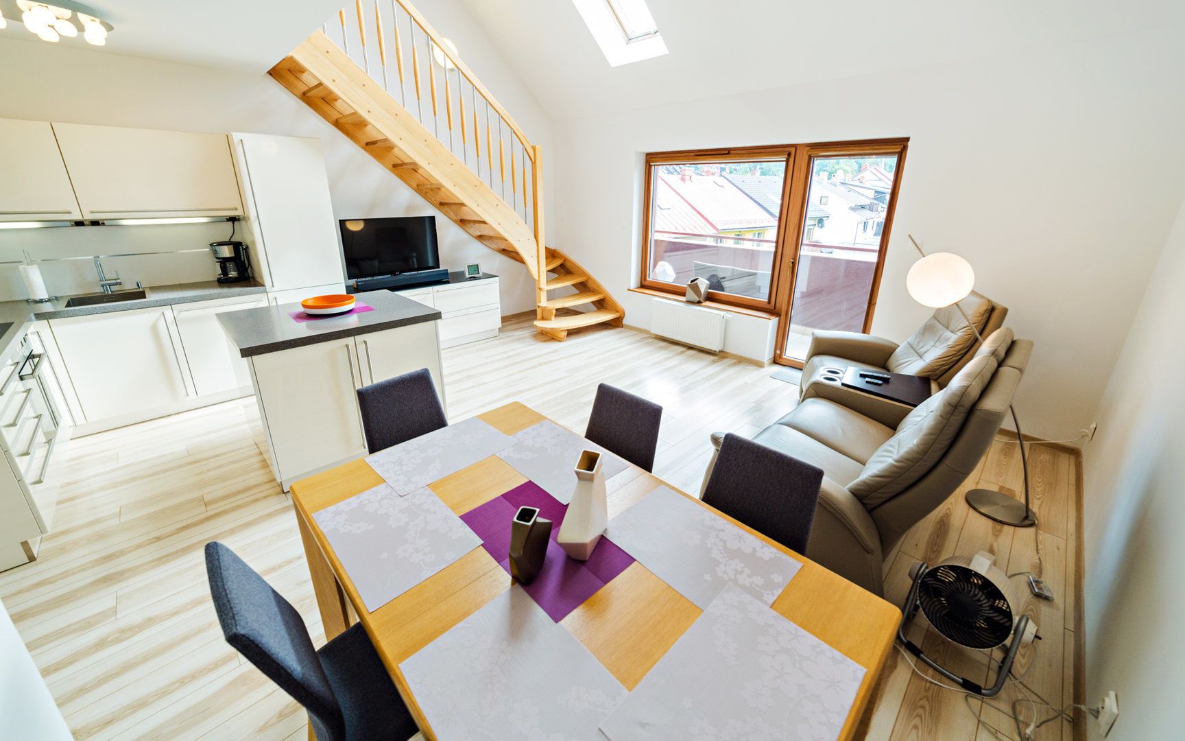 Een modern appartement met een keuken, eethoek en woonkamer. Het heeft een houten trap, een groot raam en een bank.