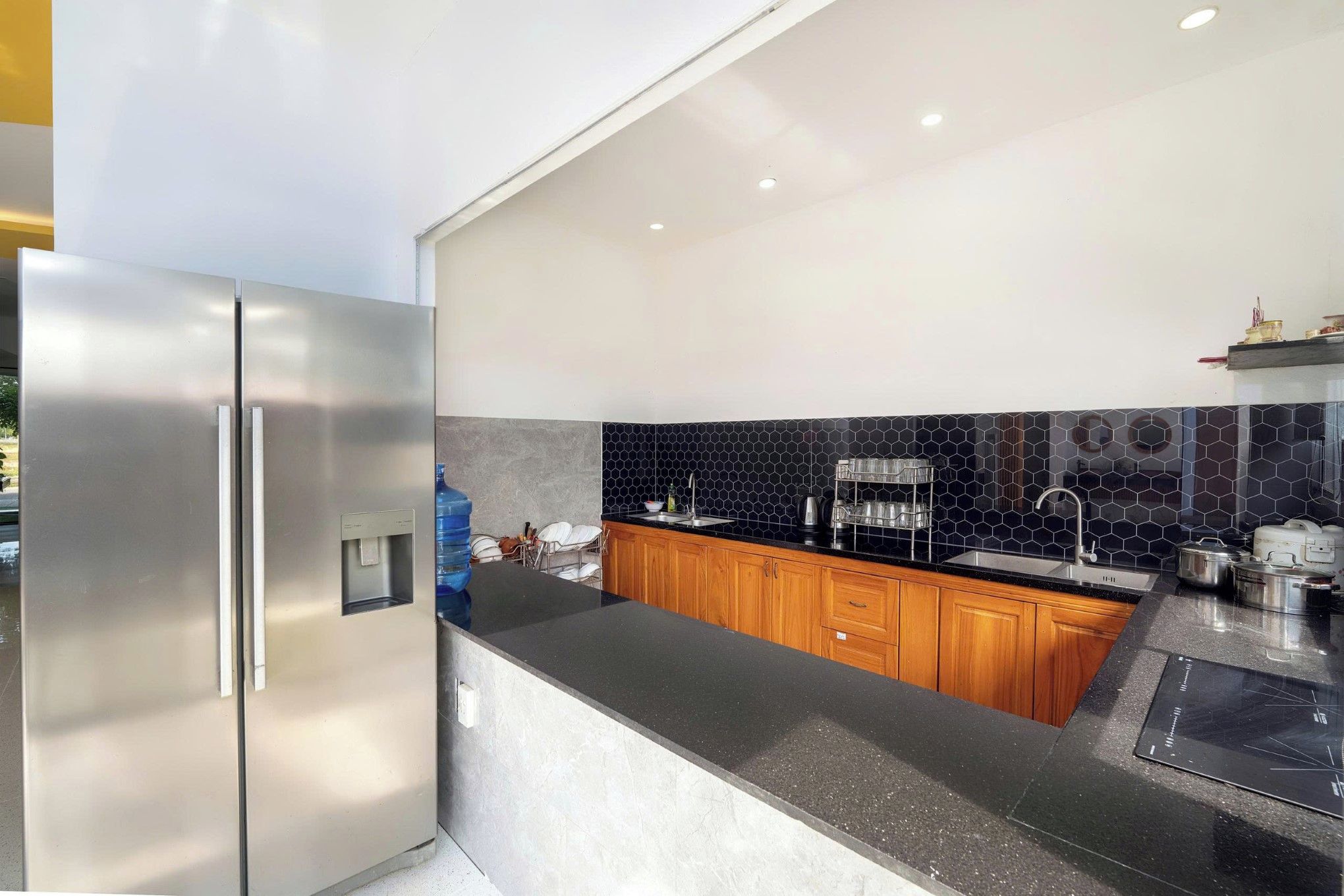 A modern kitchen with a stainless steel refrigerator, wooden cabinets, a black countertop, and a black hexagonal tile backsplash.