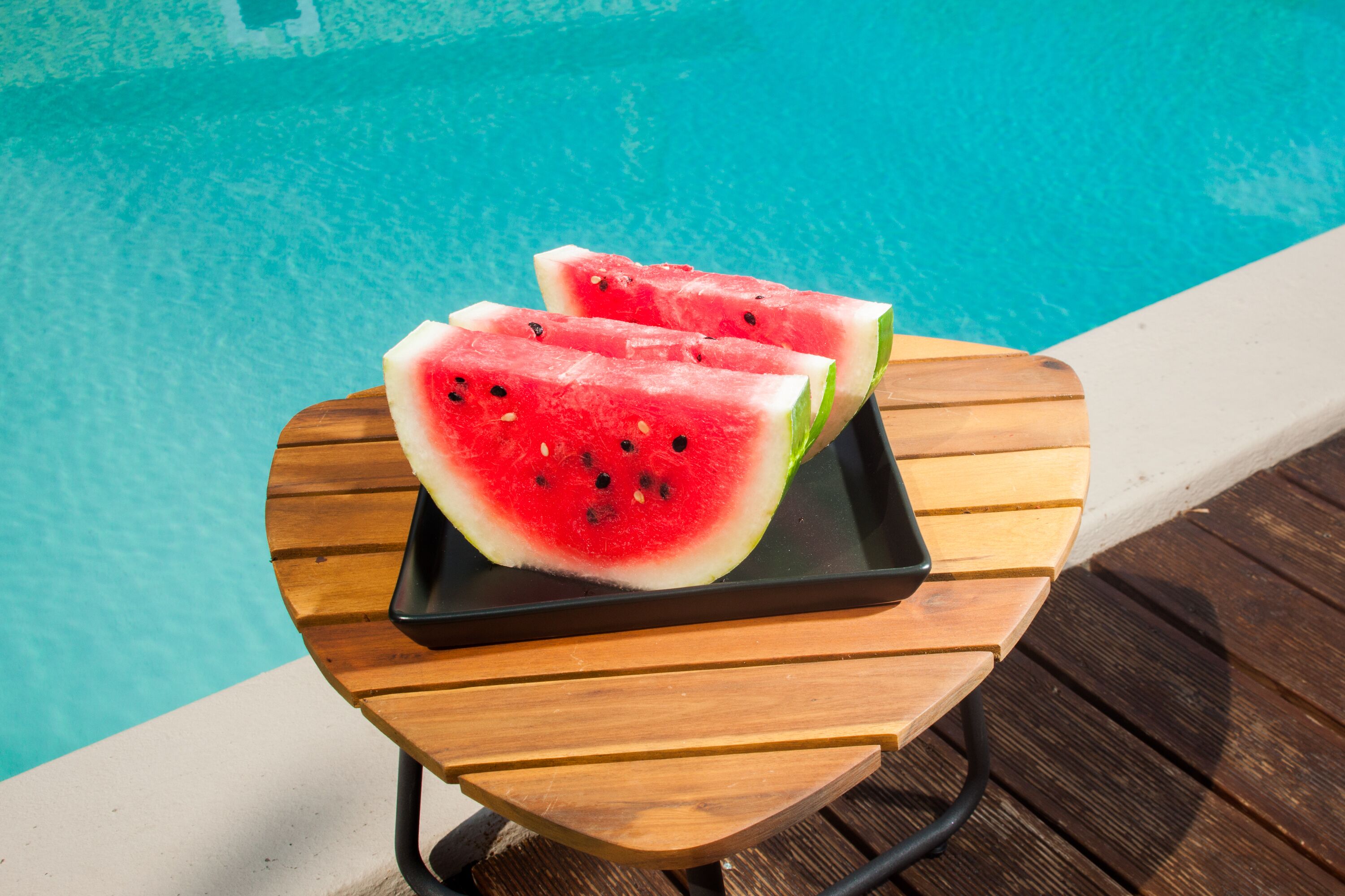 Uma mesa de madeira com uma bandeja preta exibindo duas fatias de melancia perto de uma piscina.