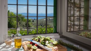 A table with a cutting board, salad, a knife, a bottle of oil, and glasses on a window sill with a view of greenery and a distant landscape. - Villas by Pelion Esties in Ano Gatzea (South Pelion)