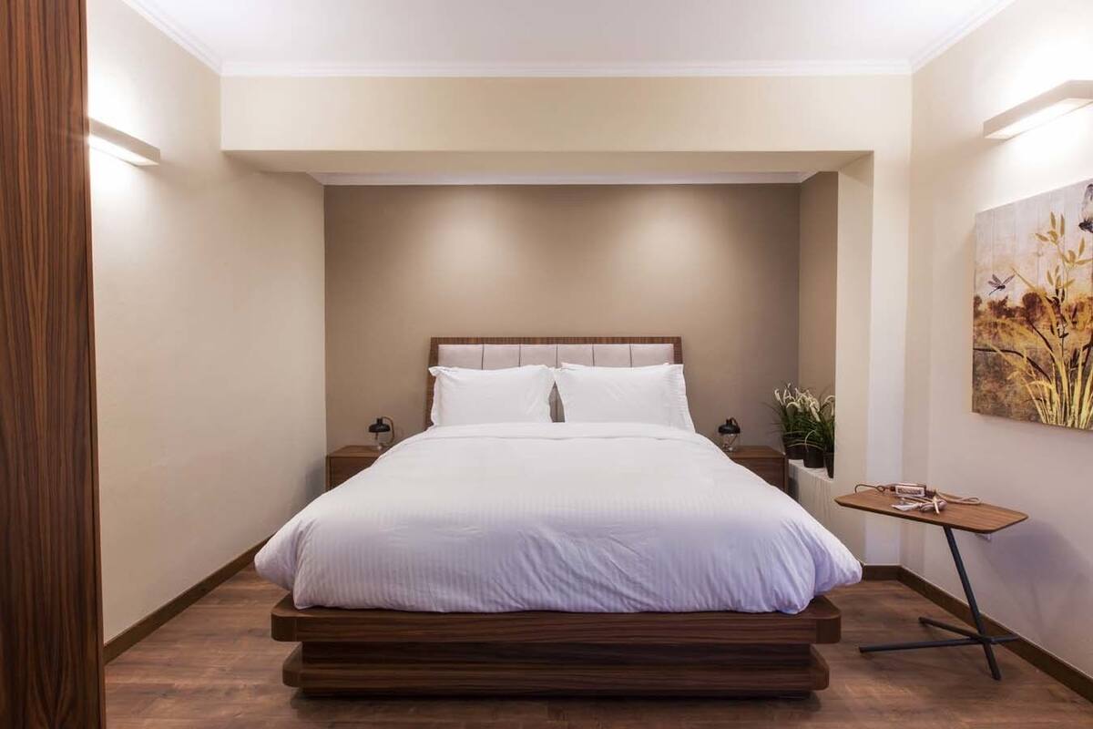 A modern bedroom with a wooden bed frame, white bedding, a nightstand with a lamp, and a small table with a plant.
