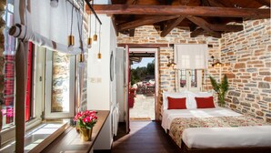 A bedroom with a stone wall, wooden beams, a bed with red pillows, a bedside table with a vase of flowers, and a view of the outside patio. - Villas by Pelion Esties in Ano Gatzea (South Pelion)