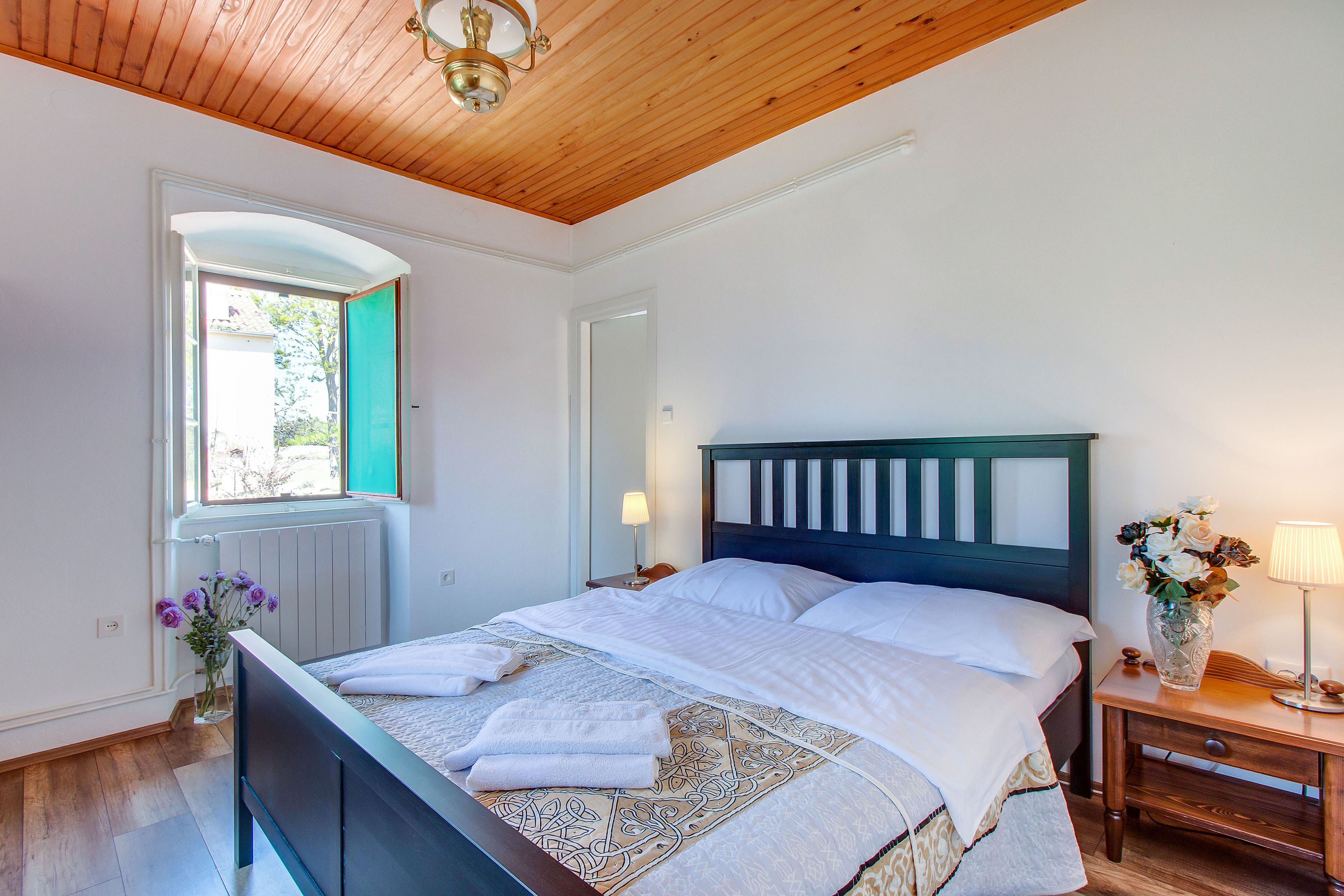 Une chambre avec un plafond en bois, un lit recouvert de draps blancs, une table de chevet avec une lampe et une fenêtre donnant sur un paysage verdoyant.