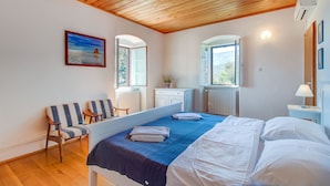 A bedroom with a wooden ceiling, a bed with white linens, a blue bedspread, a bench with striped cushions, a window with a view of the sea, and a small white cabinet. - Unique Antistress Oasis Villa Antiqua  (Mali Losinj)