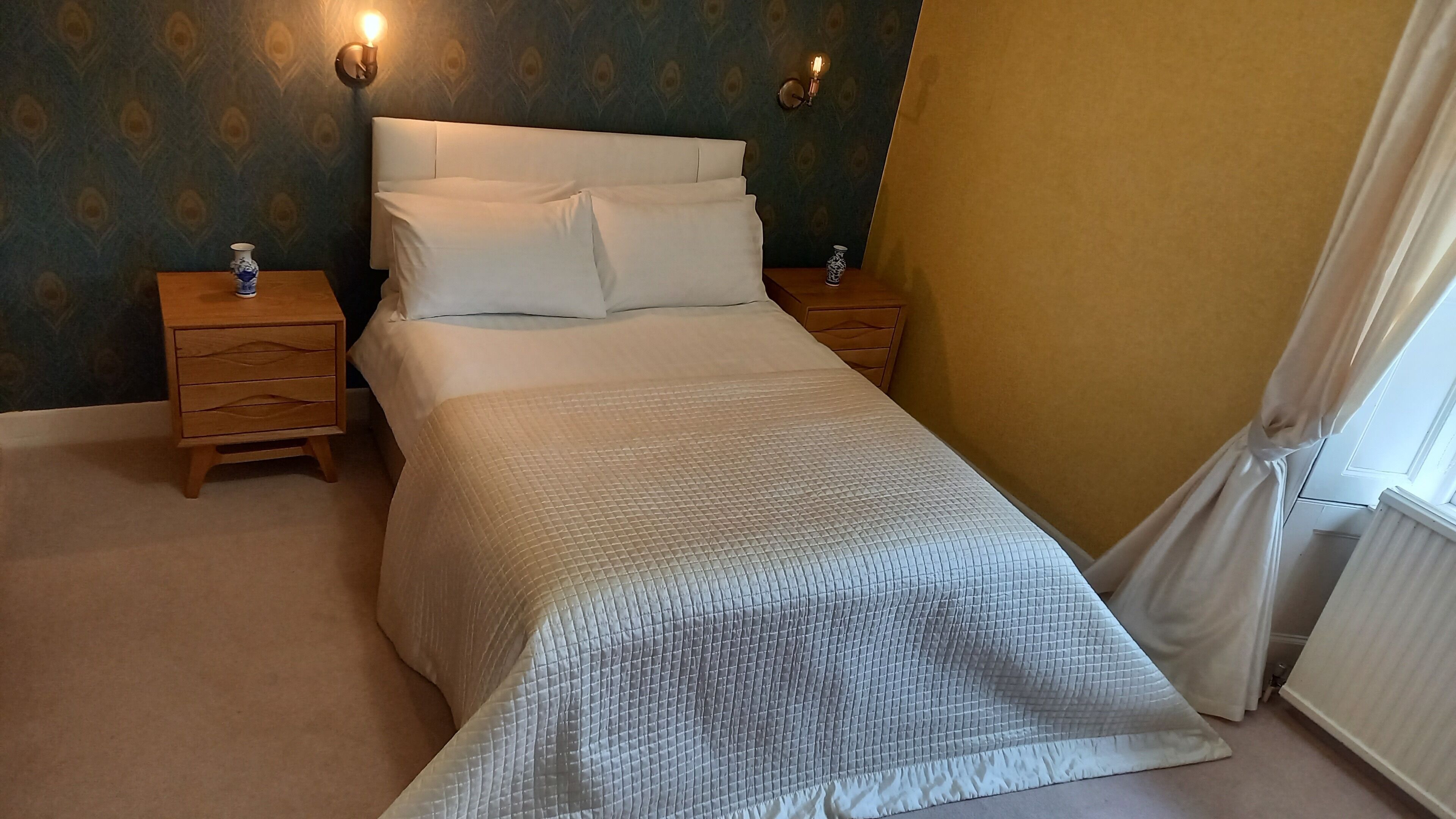 Een net opgemaakt bed met witte lakens, twee nachtkastjes met lampen, en behang met een patroon op de muren.