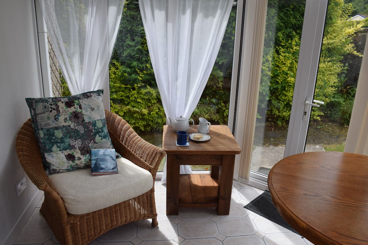 A wicker chair with a floral cushion and a book, a small wooden table with a teapot and a mug, and a view of greenery through a glass door.