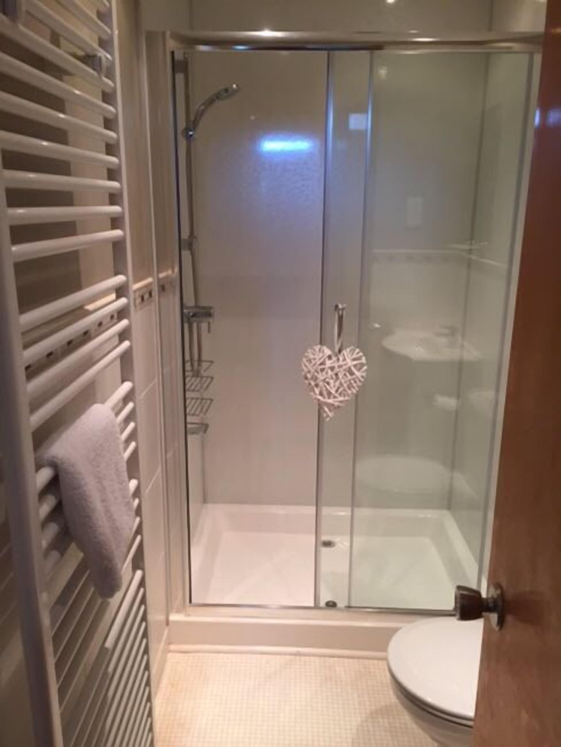 Una ducha con cerramiento de vidrio, una decoración con forma de corazón, una toalla blanca sobre un radiador y un inodoro visible a través del vidrio.