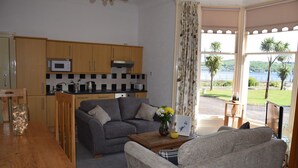 A compact living space with a kitchenette, a grey sofa, a wooden coffee table with a vase and a picture frame, and a view of a lake through large windows. - One Marine Place (Rothesay)