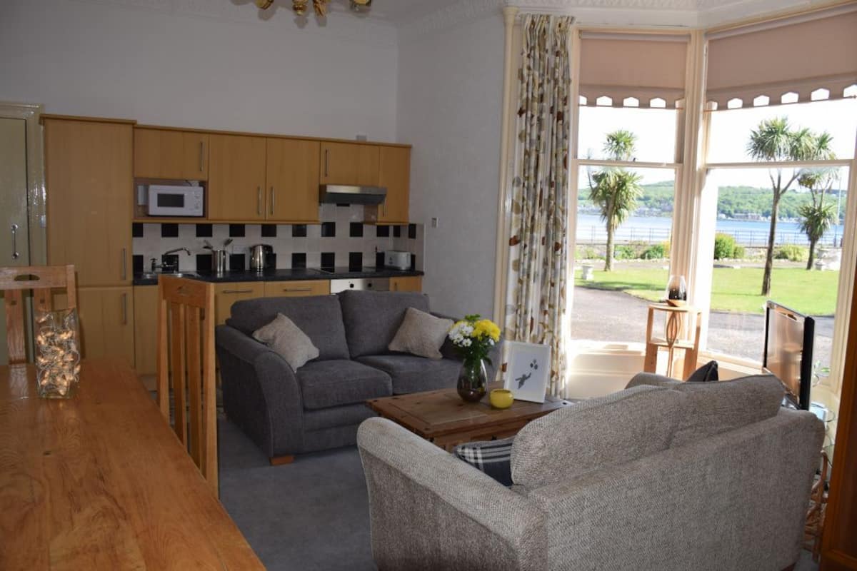 A compact living space with a kitchenette, a grey sofa, a wooden coffee table with a vase and a picture frame, and a view of a lake through large windows.