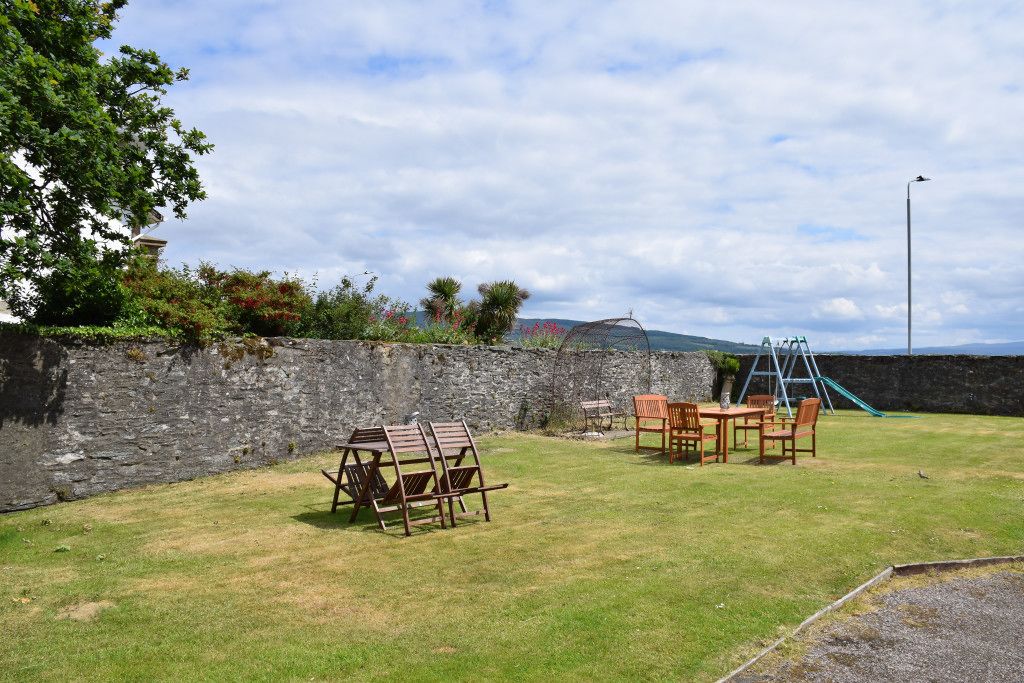 A grassy outdoor area with a stone wall, wooden chairs, a table, and a children's play structure.