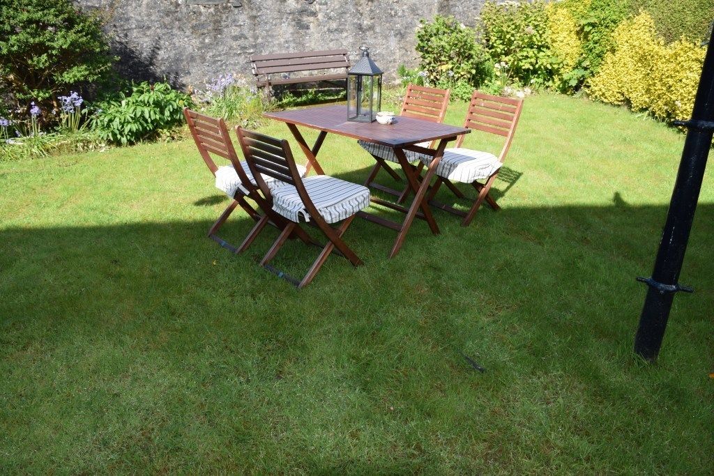 A wooden table and chairs set in a grassy garden with a stone wall in the background.