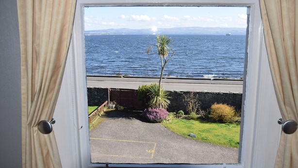 A window view of a coastal area with a clear sky, a palm tree, and a road leading to a beach. - One Marine Place (Rothesay)
