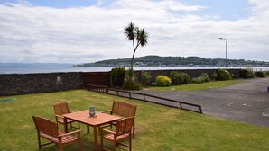 A patio with a wooden table and chairs, a palm tree, and a view of the sea and distant hills. - One Marine Place (Rothesay)
