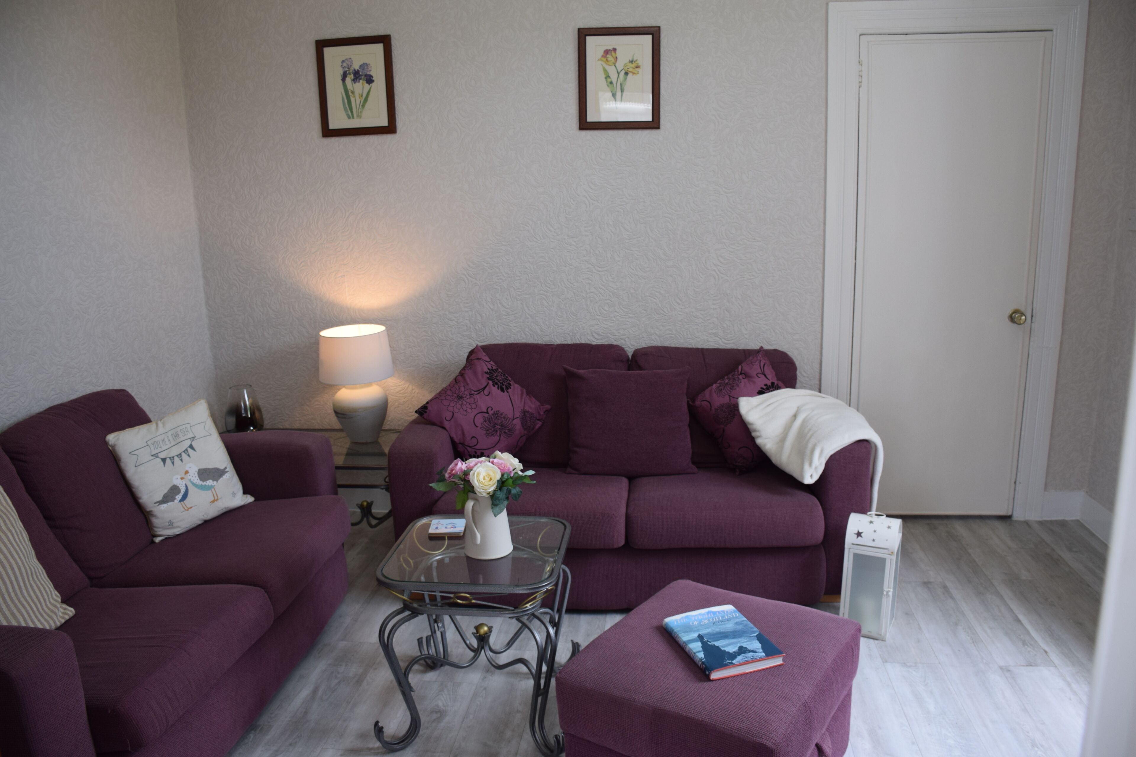 A living room with a purple sofa set, a glass coffee table with a vase of flowers, and a book on a side table.