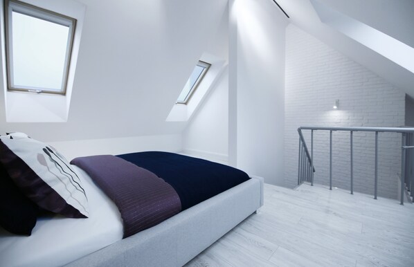 Un dormitorio moderno con techo inclinado, tragaluz, pared de ladrillo blanco y cama con cabecero.