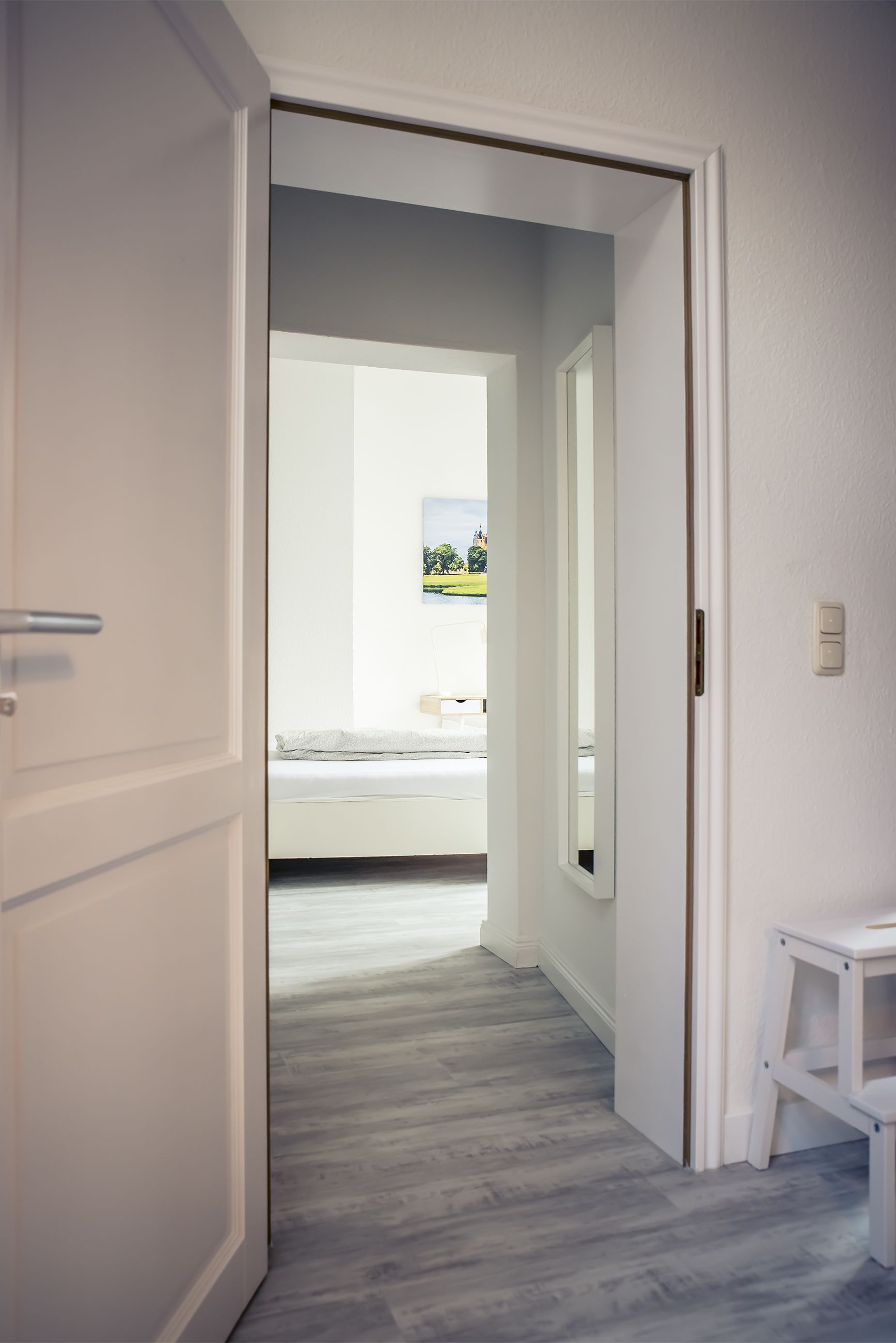 A hallway with a wooden floor leading to a bedroom with a bed and a window.