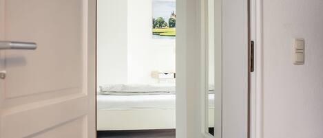 A hallway with a wooden floor leading to a bedroom with a bed and a window.