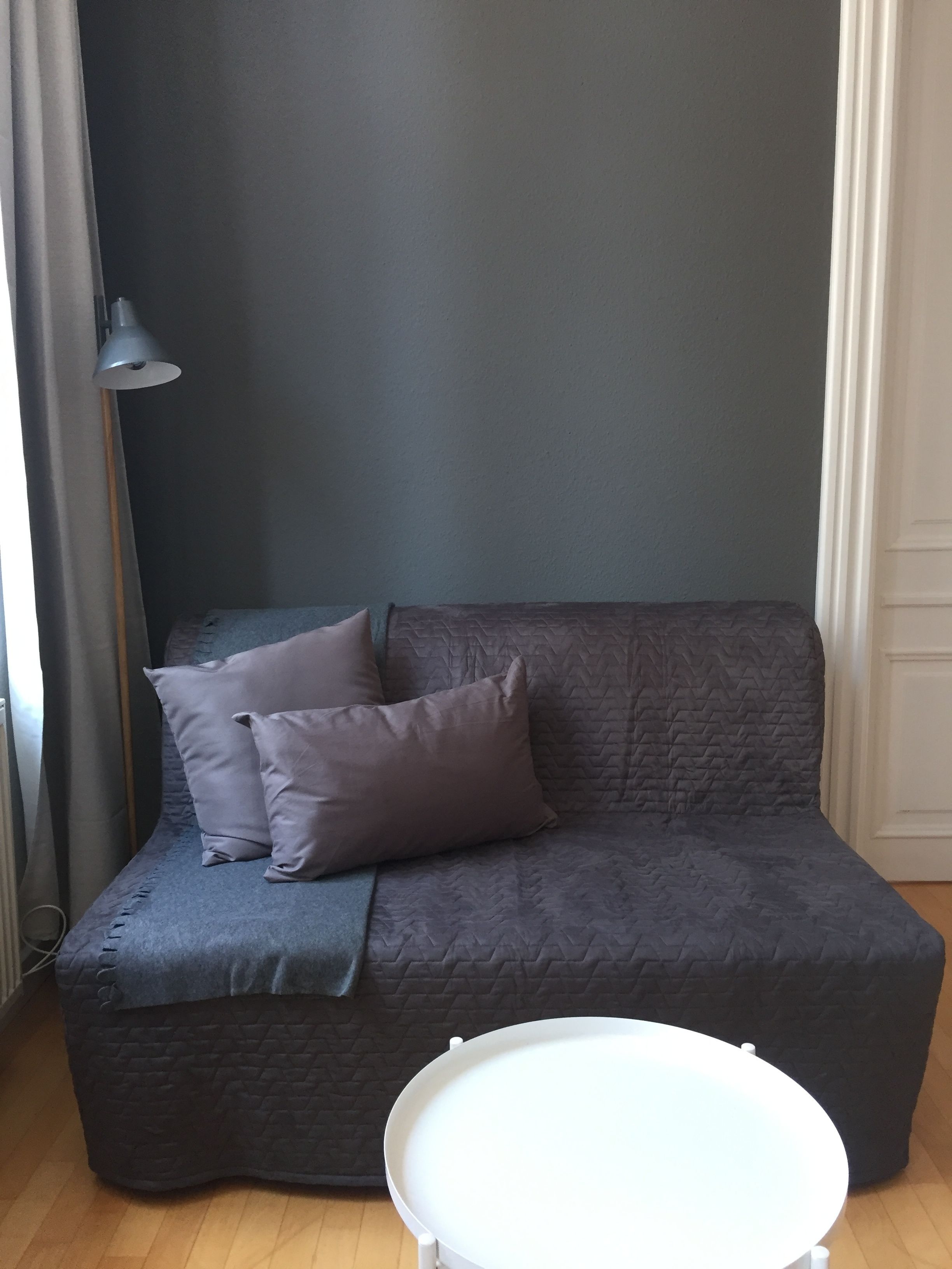 A grey sofa with a quilted texture and two pillows, a white table in front, and a grey wall.