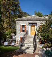 Un edificio de piedra con puerta y contraventanas de madera, una escalera que lleva a la entrada y un pequeño espacio al aire libre con sillas.