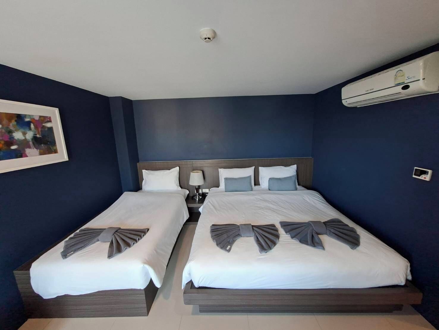 Una habitación de hotel con dos camas, aire acondicionado instalado en el techo, una mesita de noche con lámpara y un cuadro en la pared.