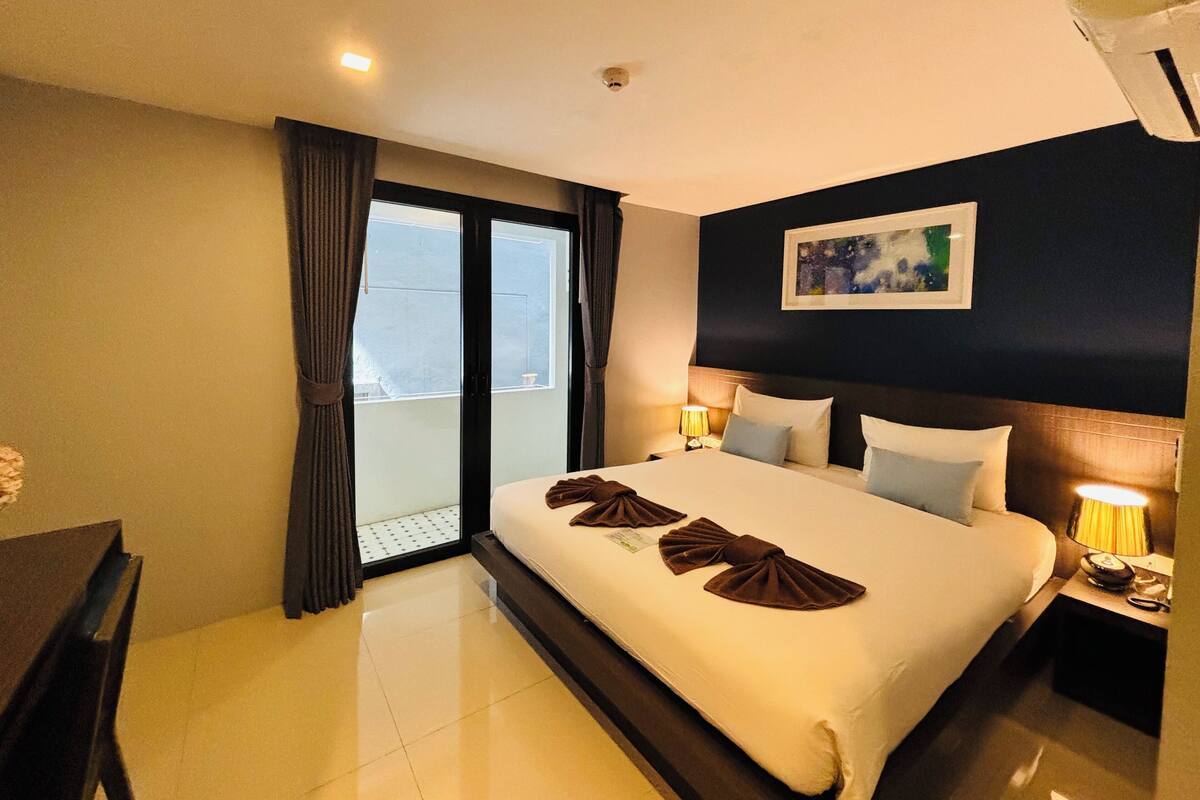 A hotel room with a large bed, two bedside tables with lamps, a window with curtains, and a painting on the wall.