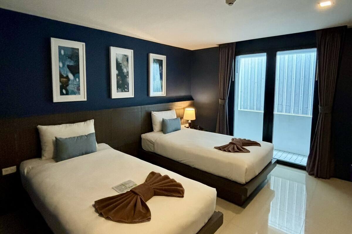 A hotel room with two beds, a nightstand with a lamp, a ceiling-mounted security camera, and a large window with curtains.
