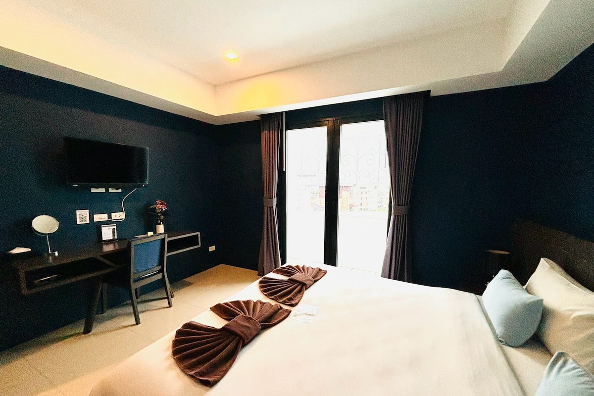 A hotel room with a bed, a desk with a computer, a TV mounted on the wall, and a window with curtains.