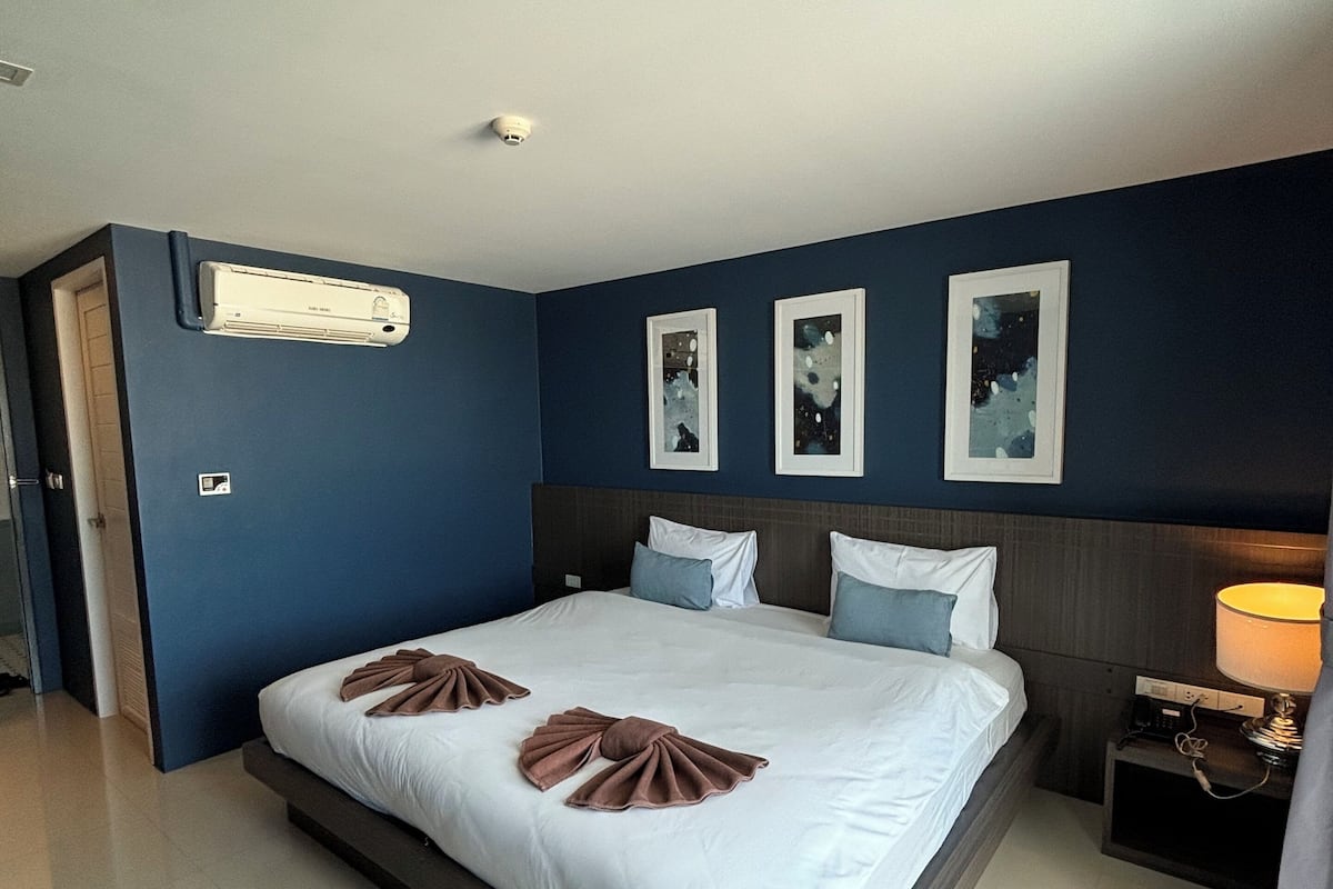 A modern hotel room with a large bed, dark blue walls, and a ceiling-mounted air conditioner.