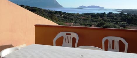 A balcony with a table and chairs, overlooking a coastal landscape with mountains and the sea.
