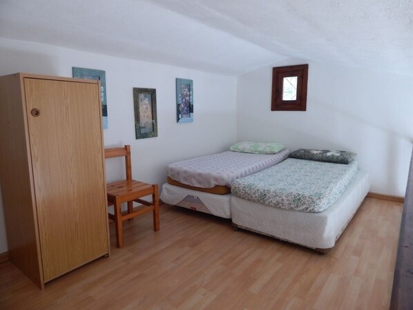 A small room with a wooden wardrobe, a single bed with a patterned bedspread, a small wooden chair, and two framed pictures on the wall.