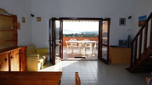 A room with a wooden bench, a yellow sofa, and a view of an outdoor patio with white chairs and a table. - AffittaSardegna - Trilo Bellavista 1 (Olbia)