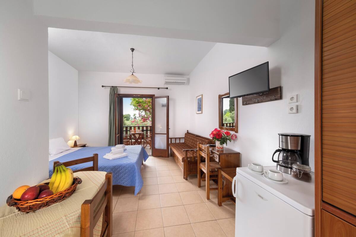 A compact hotel room with a bed, a dining table with fruit, a kitchenette, and a balcony with a view.